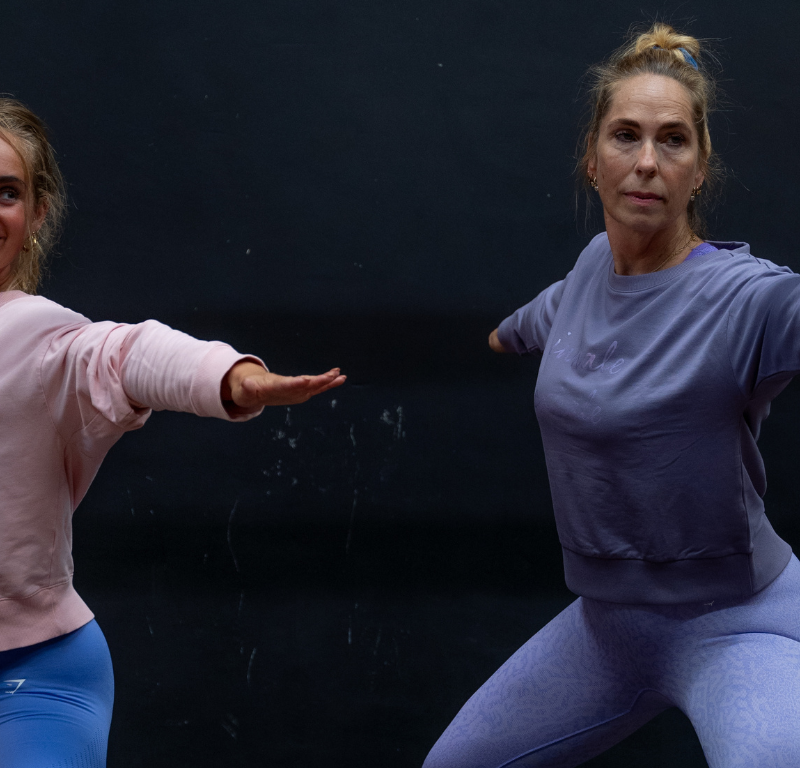 Twee vrouwen voeren samen een yogahouding uit in een sportzaal. Ze staan in een warrior pose met gestrekte armen en gefocuste blik, gekleed in sportieve kleding in pastelkleuren.